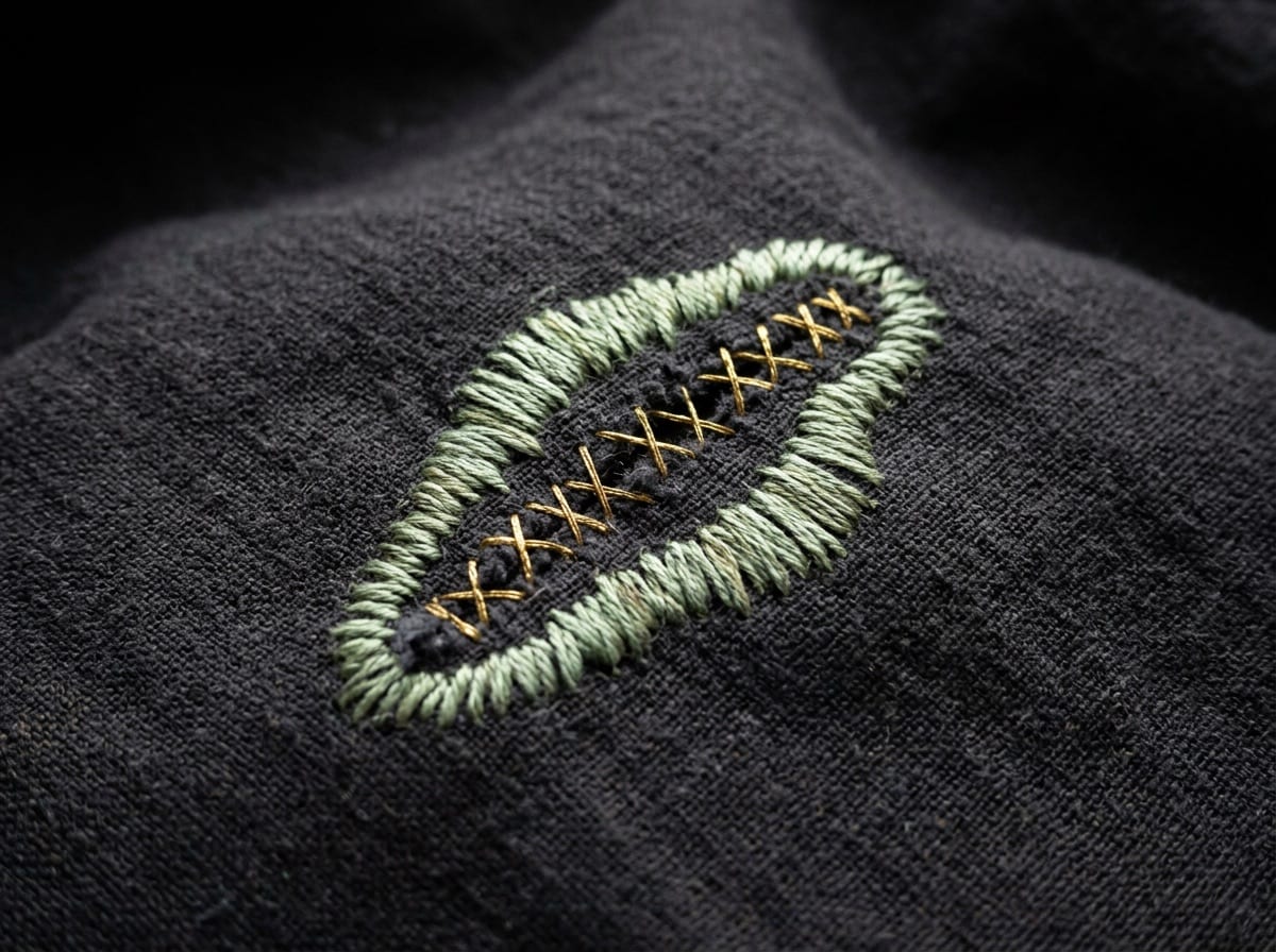 A tear in dark cloth mended with gold cross-stitch and sage satin stitch